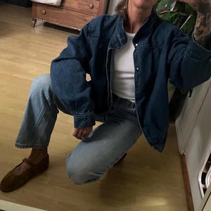 Può includere: Una persona indossa una giacca di jeans blu scuro con una t-shirt bianca sotto. Indossa anche jeans blu e scarpe di cuoio marroni.