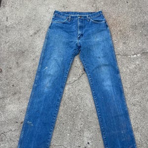 May include: Blue denim jeans with a classic five-pocket design. The jeans show signs of wear, with some discoloration and paint marks. The straight-leg cut and button-fly closure are visible.
