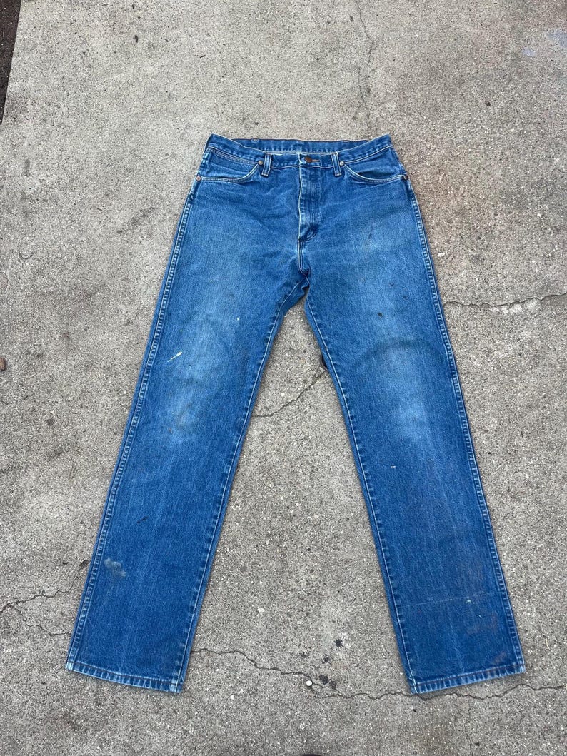 May include: Blue denim jeans with a classic five-pocket design. The jeans show signs of wear, with some discoloration and paint marks. The straight-leg cut and button-fly closure are visible.