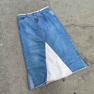 May include: A long denim skirt with a white panel sewn into the front. The skirt has a button closure and a high waist.