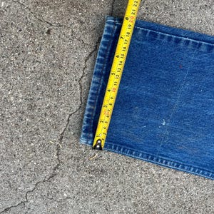 May include: A section of blue denim jeans is measured with a yellow tape measure. The tape measure shows a measurement of approximately 25 cm. The jeans are on a gray concrete surface.