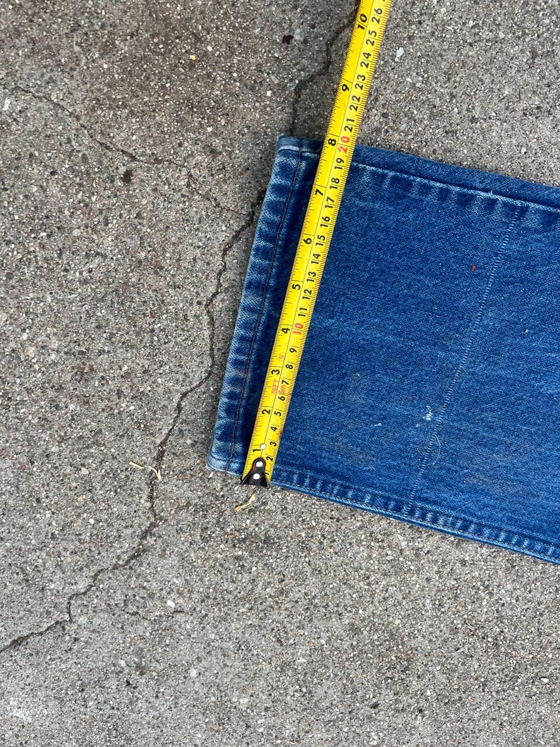 May include: A section of blue denim jeans is measured with a yellow tape measure. The tape measure shows a measurement of approximately 25 cm. The jeans are on a gray concrete surface.