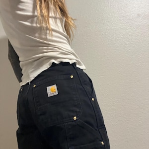 Puede incluir: Pantalones de trabajo negros con remaches dorados y un parche con el logo de Carhartt. Los pantalones tienen múltiples bolsillos y una construcción robusta. Los pantalones se combinan con una camiseta clara con dobladillo con volantes.