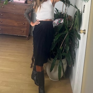 Könnte beinhalten: Eine Frau trägt ein weißes Tanktop und einen schwarzen Rock mit braunem Lederbesatz. Sie trägt schwarze Stiefel mit braunem Lederbesatz. Die Frau steht vor einer Pflanze.