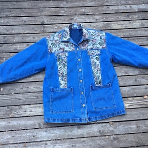 Op de afbeelding: Een blauwe spijkerjas met bloemenstofaccenten op de kraag, borst en zakken. De jas heeft een knoopsluiting en twee voorzakken. De bloemenstof heeft een mix van roze, groene en witte bloemen en bladeren.