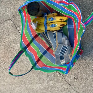 May include: A colorful striped tote bag with green, red, blue, and white stripes. Inside the bag are bananas, a stainless steel tumbler, and a folded blue and white striped cloth. The bag has green, pink, and blue handles.