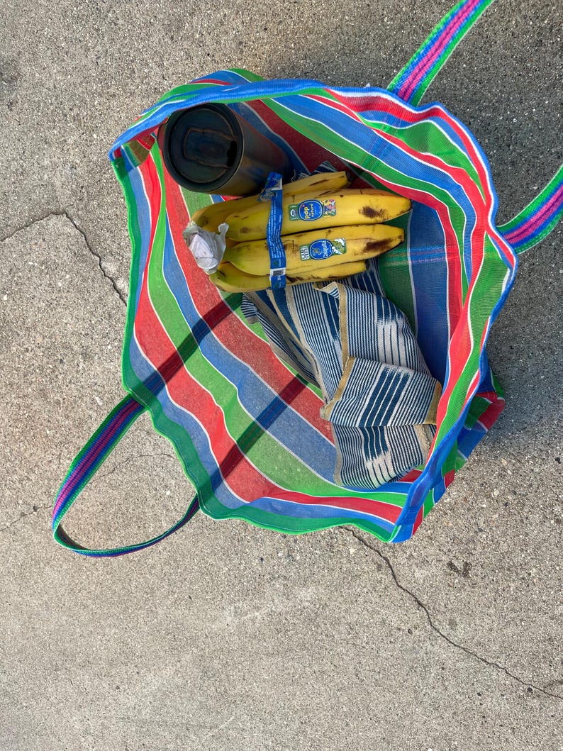 May include: A colorful striped tote bag with green, red, blue, and white stripes. Inside the bag are bananas, a stainless steel tumbler, and a folded blue and white striped cloth. The bag has green, pink, and blue handles.