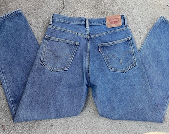 May include: Blue denim jeans with a classic five-pocket design. The jeans feature a faded wash and a leather patch with the brand logo on the back. The jeans are laid flat on a concrete surface.