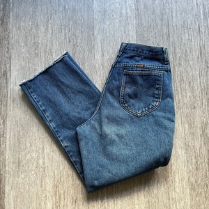 Könnte beinhalten: Eine blaue Jeanshose mit hoher Taille und geradem Bein. Die Jeans hat ein klassisches Fünf-Taschen-Design und einen ausgewaschenen Look.