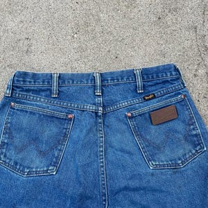 May include: Blue denim jeans featuring the Wrangler logo on the back pocket and a brown leather patch. The jeans have a classic five-pocket design and a straight leg fit.