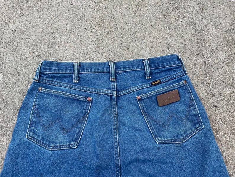 May include: Blue denim jeans featuring the Wrangler logo on the back pocket and a brown leather patch. The jeans have a classic five-pocket design and a straight leg fit.
