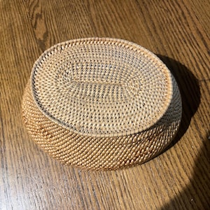 May include: A woven basket with a lid. The basket is oval-shaped and made of light brown, natural-colored woven material. The lid is also woven and fits snugly on top. The basket is sitting on a wooden surface.