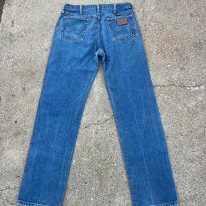 May include: Rear view of blue denim jeans with a straight leg. The jeans feature two back pockets and a brown leather patch with a logo. The jeans are made from medium-wash denim fabric.
