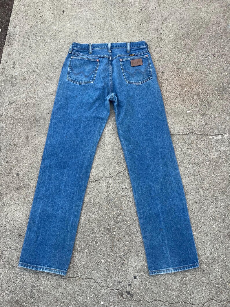May include: Rear view of blue denim jeans with a straight leg. The jeans feature two back pockets and a brown leather patch with a logo. The jeans are made from medium-wash denim fabric.
