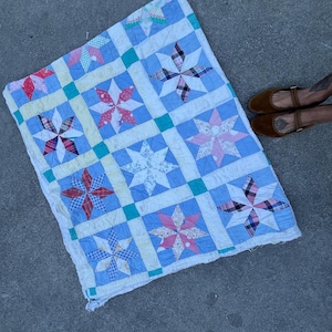 May include: A blue and white patchwork quilt with a star pattern. The quilt is made of different fabrics and colors, including pink, red, and green. The quilt is laid out on a gray concrete surface.