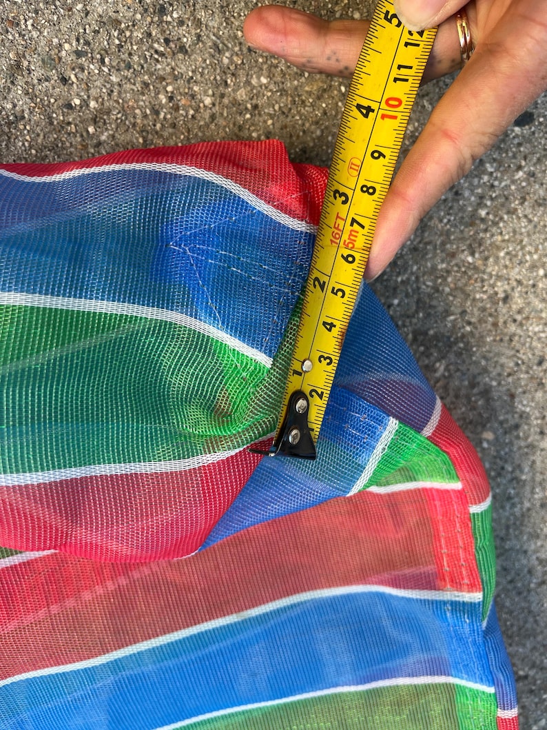 May include: Close-up of a colorful, striped mesh fabric in red, blue, green, and white. A yellow tape measure shows a measurement of approximately 4 inches. The fabric appears to be part of a larger item.