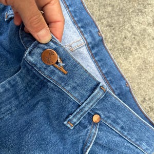 Puede incluir: Primer plano de la cintura de un par de pantalones vaqueros de mezclilla azul. Los vaqueros tienen un bot&oacute;n de cobre y un remache de cobre. La cintura est&aacute; doblada y las costuras son visibles.