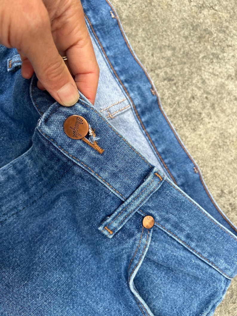 Puede incluir: Primer plano de la cintura de un par de pantalones vaqueros de mezclilla azul. Los vaqueros tienen un bot&oacute;n de cobre y un remache de cobre. La cintura est&aacute; doblada y las costuras son visibles.