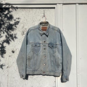 Könnte beinhalten: Eine hellblaue Jeansjacke mit Knopfleiste und zwei Fronttaschen. Die Jacke hat einen ausgewaschenen Look und hängt an einem Kleiderbügel.