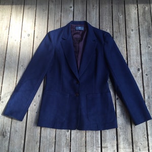 May include: A navy blue blazer with a two-button fastening and two front pockets. The blazer features a notched lapel and a dark purple lining. The garment is displayed on a weathered wooden surface.