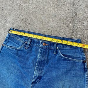 May include: Close-up of blue denim jeans with a yellow measuring tape across the waistband. The tape shows a measurement of approximately 48 cm. The jeans have classic five-pocket styling and a button and zip closure.