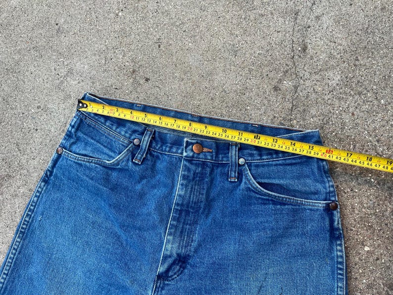 May include: Close-up of blue denim jeans with a yellow measuring tape across the waistband. The tape shows a measurement of approximately 48 cm. The jeans have classic five-pocket styling and a button and zip closure.