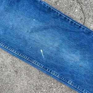 May include: Close-up of blue denim jeans showing signs of wear. The jeans have faded patches and small white paint marks. The seams are visible, and the fabric has a textured appearance.