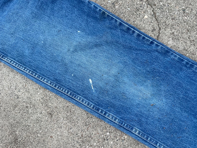 May include: Close-up of blue denim jeans showing signs of wear. The jeans have faded patches and small white paint marks. The seams are visible, and the fabric has a textured appearance.
