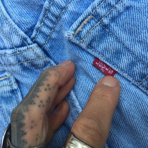 Puede incluir: Primer plano de una tela vaquera azul claro con una etiqueta roja de Levi's cosida en la tela. La etiqueta dice "Levi's" y tiene una peque&ntilde;a pesta&ntilde;a roja con el n&uacute;mero "502" en ella.