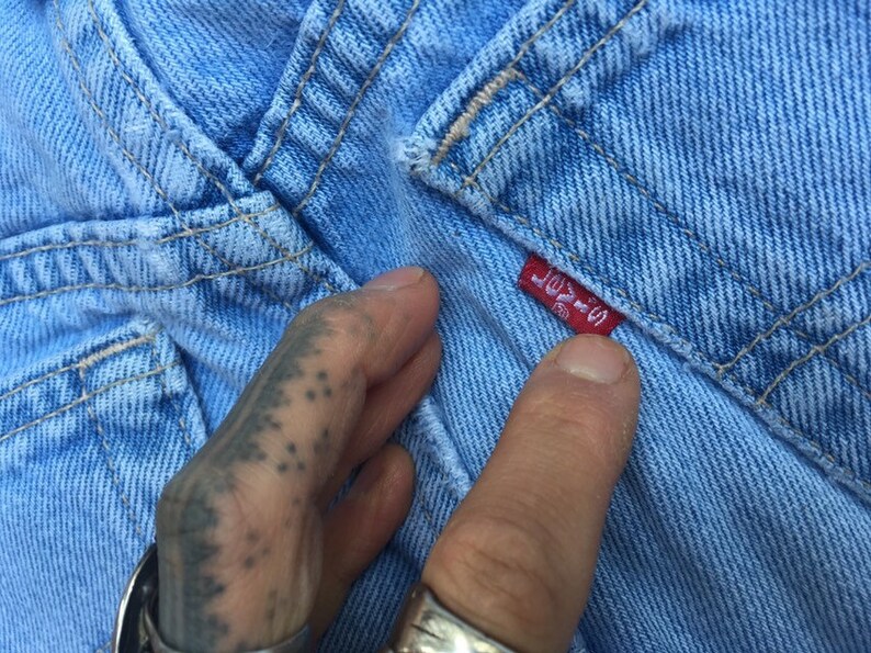 Puede incluir: Primer plano de una tela vaquera azul claro con una etiqueta roja de Levi's cosida en la tela. La etiqueta dice "Levi's" y tiene una peque&ntilde;a pesta&ntilde;a roja con el n&uacute;mero "502" en ella.