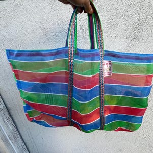 May include: A colorful, striped tote bag with blue, green, red, and orange horizontal stripes. The bag has braided handles in matching colors and a small tag attached. The bag is held up against a white wall.
