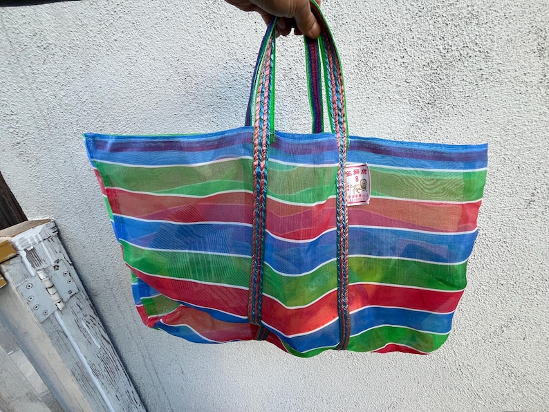 May include: A colorful, striped tote bag with blue, green, red, and orange horizontal stripes. The bag has braided handles in matching colors and a small tag attached. The bag is held up against a white wall.