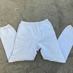 Könnte beinhalten: Hellgraue Jogginghose mit elastischem Bund und Kordelzug. Die Hose hat elastische Bündchen an den Knöcheln. Das Kleidungsstück ist aus einem weichen, bequemen Material gefertigt. Die Jogginghose liegt auf einer Betonoberfläche.