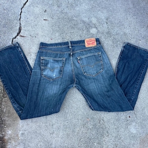 May include: A white ceramic mug with a black handle and the text "But First, Coffee" in a bold, black font. The mug is filled with a dark liquid, likely coffee, and sits on a light-colored surface.