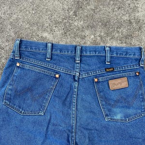 Puede incluir: Un par de pantalones vaqueros azules de mezclilla con un parche de cuero marr&oacute;n que dice "Wrangler" en el bolsillo trasero.