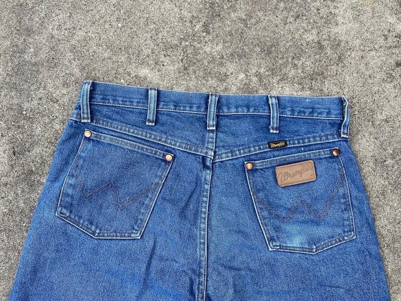 Puede incluir: Un par de pantalones vaqueros azules de mezclilla con un parche de cuero marr&oacute;n que dice "Wrangler" en el bolsillo trasero.