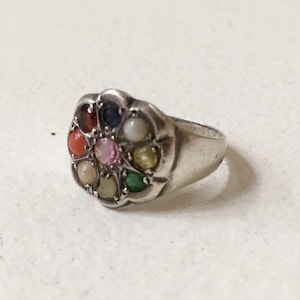 May include: A silver ring with a flower-shaped setting featuring nine colorful gemstones. The ring is set with red, orange, yellow, green, blue, purple, and white stones.