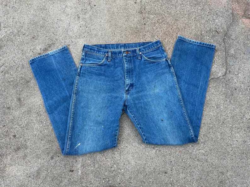 May include: Blue denim jeans with a classic five-pocket design. The jeans have a button and zip closure, belt loops, and a straight leg cut. The denim shows some wear and tear, with a few paint marks. A casual, everyday wardrobe staple.