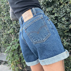 Puede incluir: Un par de pantalones cortos de mezclilla azul con dobladillo. Los pantalones cortos tienen un bolsillo trasero con una etiqueta de la marca Levi's.