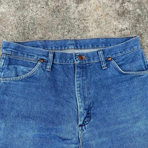 Puede incluir: Un par de pantalones vaqueros azules de mezclilla con un estilo cl&aacute;sico de cinco bolsillos. Los vaqueros tienen una pierna recta y una bragueta con botones.