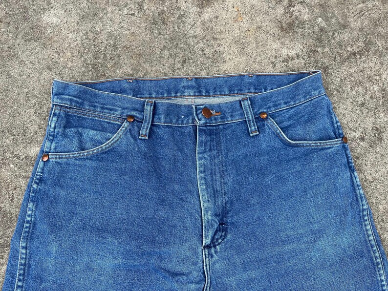 Puede incluir: Un par de pantalones vaqueros azules de mezclilla con un estilo cl&aacute;sico de cinco bolsillos. Los vaqueros tienen una pierna recta y una bragueta con botones.