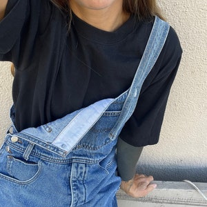 May include: A person wearing a black t-shirt and blue denim dungarees. The dungarees have a white fabric strap and a button closure.
