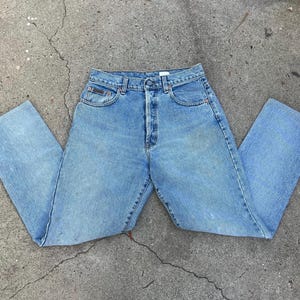Puede incluir: Vaqueros de mezclilla azul claro con un diseño clásico de cinco bolsillos. Los vaqueros tienen una cintura alta y un corte de pierna recto. El denim tiene un lavado descolorido, lo que le da un aspecto vintage. Los vaqueros están planos sobre una superficie de hormigón.