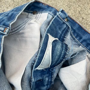 May include: Close-up view of blue denim jeans, focusing on the button closure and interior pocket lining. The jeans have a classic, worn appearance with visible stitching and a faded wash. The fabric appears to be robust and well-constructed.
