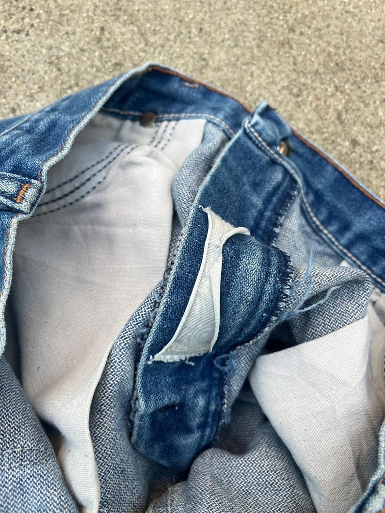 May include: Close-up view of blue denim jeans, focusing on the button closure and interior pocket lining. The jeans have a classic, worn appearance with visible stitching and a faded wash. The fabric appears to be robust and well-constructed.