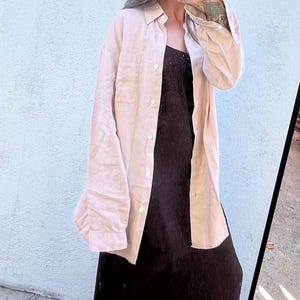 Könnte beinhalten: Ein hellbeiges, langärmeliges Hemd mit Knopfleiste, Kragen und einer Brusttasche, getragen über einem dunkelbraunen, ärmellosen Slip-Kleid. Das Hemd hat weiße Knöpfe an der vorderen Knopfleiste und an den Manschetten der langen Ärmel.