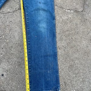 May include: Blue denim jeans with a straight leg design. A yellow measuring tape is placed along the jeans, showing measurements in inches. The jeans have some wear and tear, with visible marks on the fabric.