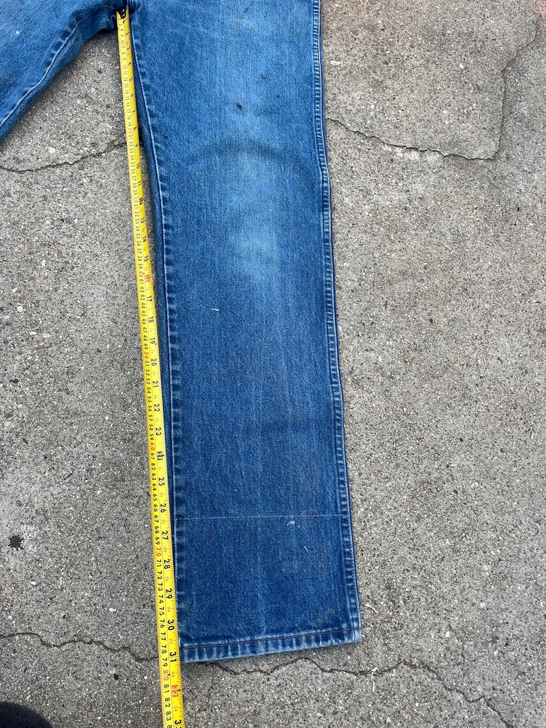 May include: Blue denim jeans with a straight leg design. A yellow measuring tape is placed along the jeans, showing measurements in inches. The jeans have some wear and tear, with visible marks on the fabric.