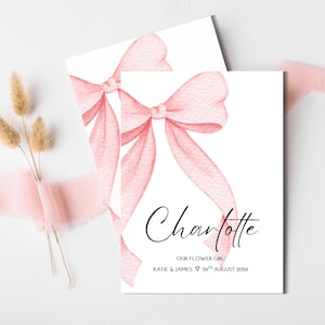 May include: A white rectangular card featuring a large watercolor pink bow illustration. The text reads Charlotte, OUR FLOWER GIRL, KATIE & JAMES 29TH AUGUST 2026. The card is accented with a soft pink frayed ribbon and dried tan grass stems.