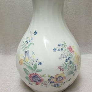 Könnte beinhalten: Eine weiße Keramikvase mit einem Blumenmuster. Die Vase hat eine breite Öffnung und einen runden Körper. Die Blumen sind in den Farben Rosa, Gelb, Blau und Grün.